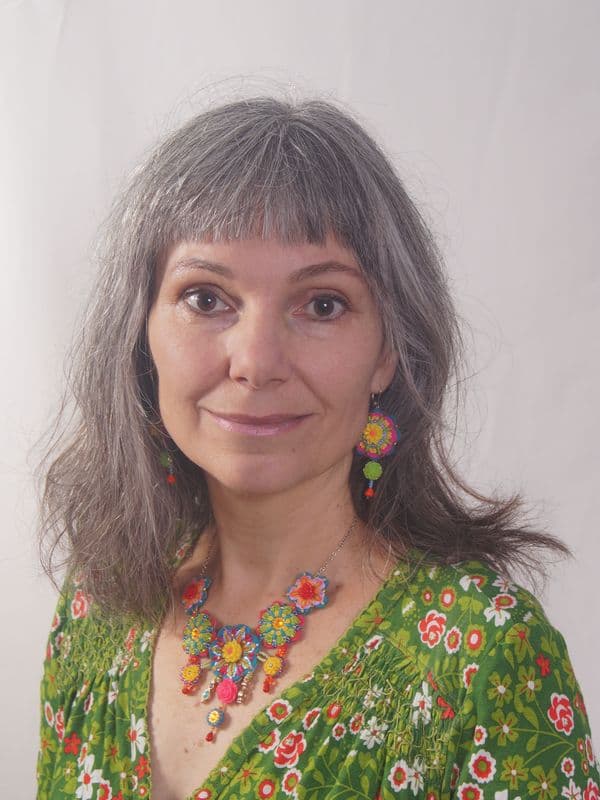 Porträt einer Frau mit grau-braunen Haaren, einem grünen Kleid mit Blumen, einer Kette sowie Ohrringen mit je gehäkelten und bestickten Blumen