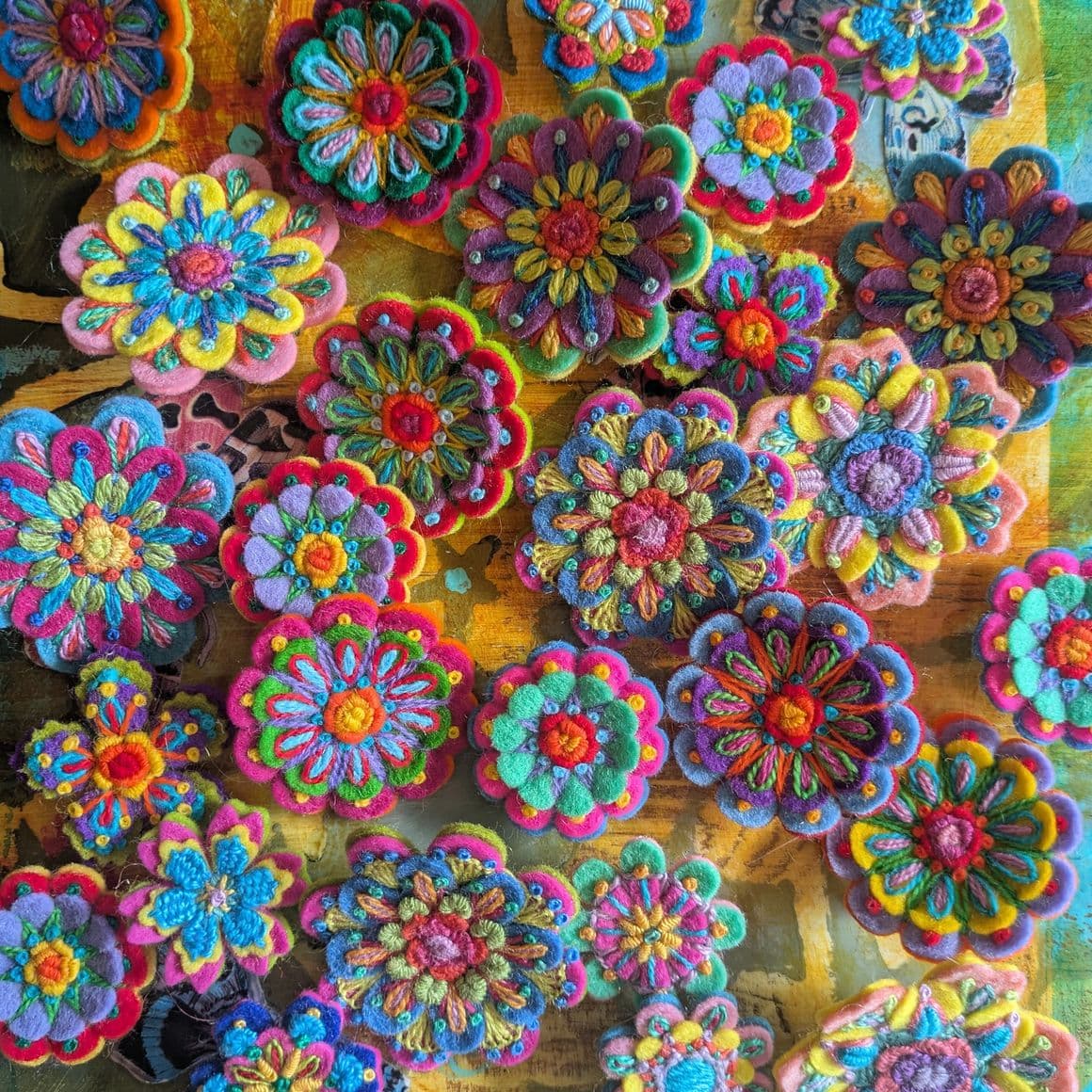 Mehrere handbestickte Filzblumen in unterschiedlichen Größen und Formen liegen dicht nebeneinander auf einer bunten Unterlage. Die Filzblumen sind in kräftigen Farben wie Blau, Pink, Gelb, Grün und Lila gestaltet und mit kontrastierenden Garnen detailreich bestickt. Jede Blume zeigt ein individuelles Muster aus konzentrischen Kreisen, Blütenblättern und Ziernähten. Die Stickereien heben die Strukturen und Farbübergänge der einzelnen Filzblumen hervor.