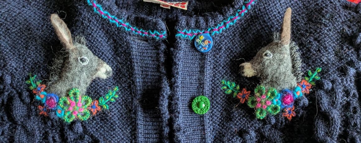 Dunkelblaue Strickjacke mit plastischer Zopfmusterstruktur. Auf beiden Seiten der Brustpartie ist eine aufwendige Handstickerei zu sehen: Jeweils ein plastisch gestalteter Eselkopf mit aufgestellten Ohren, umgeben von farbenfrohen, gestickten Blumen und Blättern. Die Knopfleiste ist mit unterschiedlich farbigen Knöpfen in Grün, Rot und Blau versehen. Am Halsausschnitt verläuft eine bunte Zierstickerei.