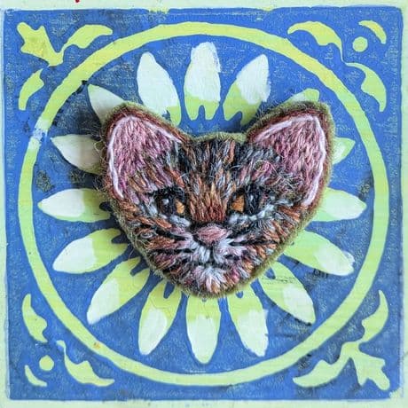 Im Mittelpunkt steht eine handgestickte Katzenbrosche. Die Brosche zeigt einen Katzenkopf mit spitzen Ohren, gestickt in verschiedenen Brauntönen mit weißen und schwarzen Akzenten. Die Brosche liegt auf einer rechteckigen Karte mit blauem Hintergrund und einem gelben, kreisförmigen Blumenmuster.
