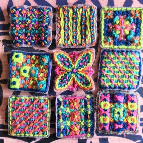 Neun handgestickte Quadrate liegen in drei Reihen angeordnet auf einer gemusterten Unterlage. Jedes Quadrat zeigt ein individuelles, farbenfrohes Stickmuster mit geometrischen und floralen Motiven. Die verwendeten Garne sind leuchtend und kontrastreich, darunter Gelb, Orange, Grün, Blau und Pink. Die Stickereien sind dicht gearbeitet und heben sich deutlich vom dunklen Untergrund der Quadrate ab.