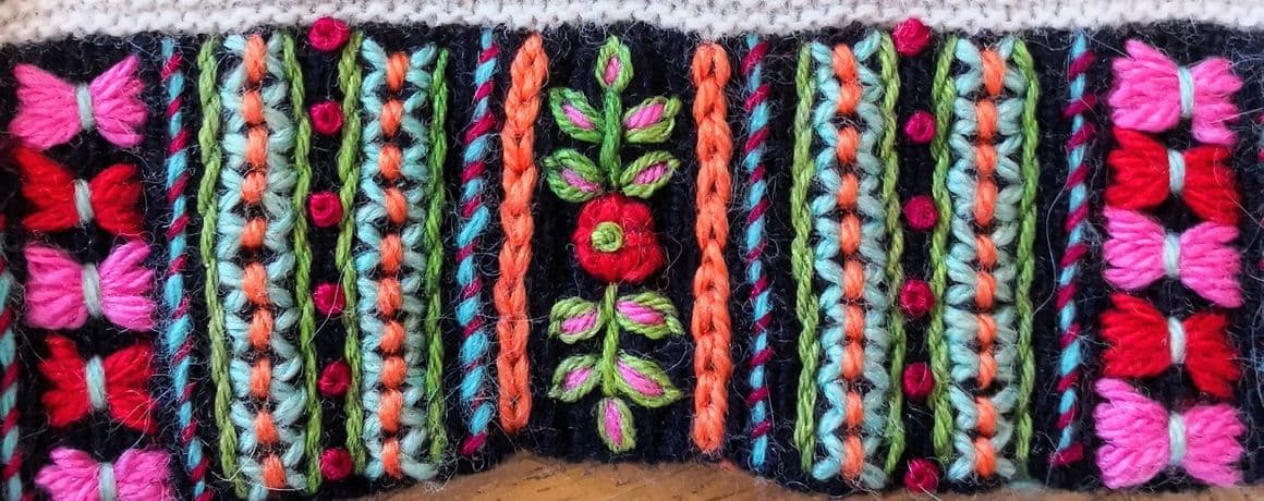 Im Vordergrund verläuft eine gestickte Borte entlang des unteren Bildrands. Die Borte zeigt ein farbenfrohes Muster mit vertikalen Streifen in Pink, Rot, Grün, Türkis und Orange. Dazwischen sind florale und geometrische Motive eingearbeitet. Darüber befindet sich ein gestrickter Stoff mit schmalen, waagerechten schwarz-weißen Streifen.