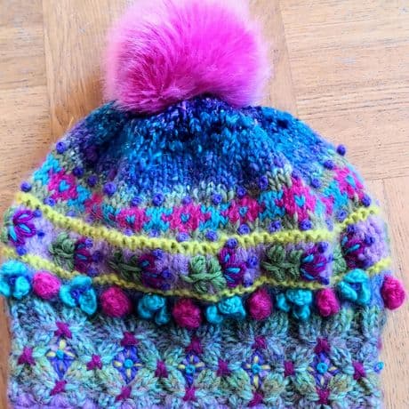 Handgestrickte Mütze mit auffälligem, mehrfarbigem Muster. Die Mütze zeigt verschiedene Stricktechniken und Farbkombinationen in Blau, Grün, Lila und Pink. Am oberen Ende befindet sich ein großer, flauschiger Bommel in kräftigem Pink. Die Mütze ist mit kleinen, eingestrickten und aufgesetzten Details wie Blüten und Noppen verziert. Der Hintergrund besteht aus einer hellen Holzoberfläche.