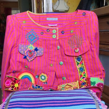 Eine handbestickte Jacke mit leuchtend pink-orangen Streifen liegt auf einer gestreiften Unterlage. Die Vorderseite ist mit zahlreichen farbenfrohen Stickereien verziert, darunter geometrische Muster, ein stilisiertes Herz mit Flammen, ein Regenbogen, Blumenmotive und verschiedene Bänder mit Mustern. Die Jacke hat einen runden Halsausschnitt und zwei Brusttaschen. Die Stickereien sind detailreich und in kräftigen Farben ausgeführt.