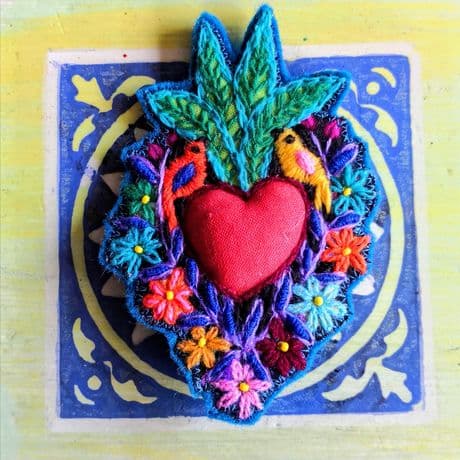 Brosche in Herzform mit rotem, textilen Mittelteil, umgeben von farbenfrohen, gestickten Blumen und zwei kleinen Vögeln. Über dem Herz sind grüne, gestickte Blätter angeordnet. Die Brosche liegt auf einer blau-weißen Fliese mit floralem Muster auf gelbem Untergrund.