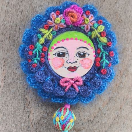 Runde Brosche mit gehäkeltem Rand in kräftigem Blau, verziert mit bunten Stickereien in Rot, Grün, Gelb, Rosa und Orange.In der Mitte ist ein rundes handbemaltes Gesicht zu sehen. Im oberen Bereich sind florale Muster eingearbeitet, darunter eine kleine, mehrfarbige Kugel als Anhänger. 
