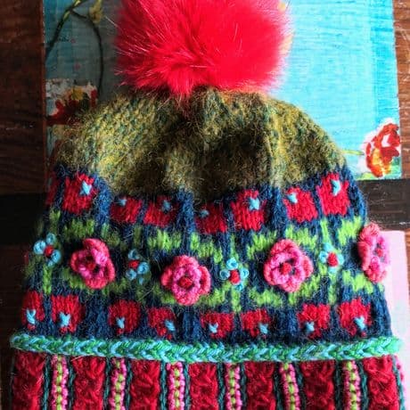 Runde Brosche mit gehäkeltem Rand in kräftigem Blau, verziert mit bunten Stickereien in Rot, Grün, Gelb, Rosa und Orange. Im oberen Bereich sind florale Muster eingearbeitet, darunter eine kleine, mehrfarbige Kugel als Anhänger. Die Brosche liegt auf einer Holzoberfläche.Handgestrickte Mütze mit auffälligem, farbenfrohem Muster. Der untere Rand zeigt ein breites, rot-blau-grünes Zopfmuster. Darüber verläuft ein florales Muster mit eingestrickten und aufgesetzten rosa Blüten sowie kleinen blauen Punkten. Die Hauptfläche ist olivgrün mit roten und blauen Akzenten. Oben sitzt ein großer, flauschiger Bommel in kräftigem Rot. 